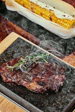 Mayalanmış biftek. Siyah bir tabakta biberiyeli biftek ve yeşil, baharatlı ve taze sebzeli ızgara mısır. Bakır bir çarşaf üzerinde. Kapat. seri fotoğraf
