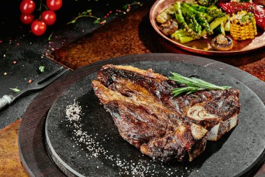 Izgara sebzelerle servis edilen T-Bone bifteği siyah yuvarlak bir tabakta yeşil, baharat ve sebzelerden oluşan bir arka planda, bakır bir çarşaf üzerinde. Kapat.