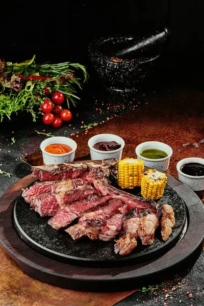 T-Bone Steak podávaný s grilovanou zeleninou a různými omáčkami na černém kulatém talíři na pozadí zeleně, koření a zeleniny, na měděném plechu. zavřít — Stock fotografie