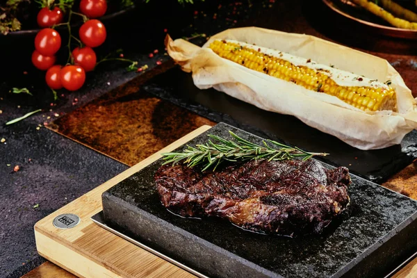 Kvašený steak. Steak s rozmarýnem na černém talíři podávaný s grilovanou kukuřicí na pozadí zeleně, koření a čerstvé zeleniny, na měděném plechu. Zblízka. série fotografií — Stock fotografie