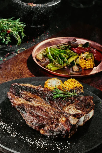 T-Bone steak podávaný s grilovanou zeleninou na černém kulatém talíři na pozadí zeleně, koření a zeleniny, na měděném plechu. zavřít — Stock fotografie