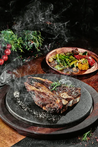 T-Bone steak podávaný s grilovanou zeleninou na černém kulatém talíři na pozadí zeleně, koření a zeleniny, na měděném plechu. zavřít — Stock fotografie