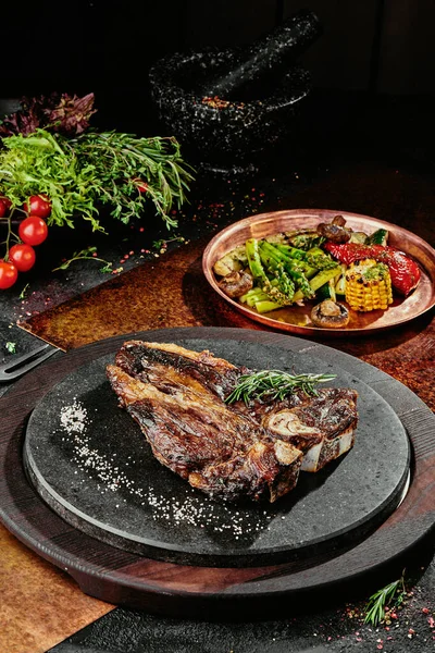 T-Bone steak podávaný s grilovanou zeleninou na černém kulatém talíři na pozadí zeleně, koření a zeleniny, na měděném plechu. zavřít — Stock fotografie