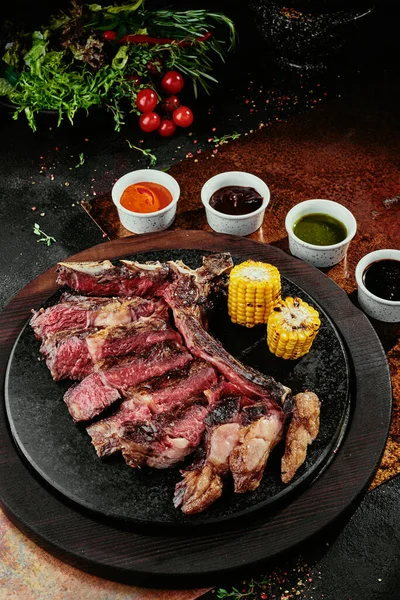 T-Bone Steak podávaný s grilovanou zeleninou a různými omáčkami na černém kulatém talíři na pozadí zeleně, koření a zeleniny, na měděném plechu. zavřít — Stock fotografie