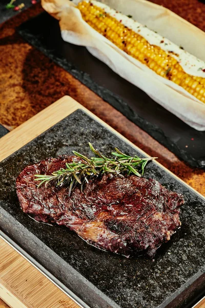 Kvašený steak. Steak s rozmarýnem na černém talíři podávaný s grilovanou kukuřicí na pozadí zeleně, koření a čerstvé zeleniny, na měděném plechu. Zblízka. série fotografií — Stock fotografie