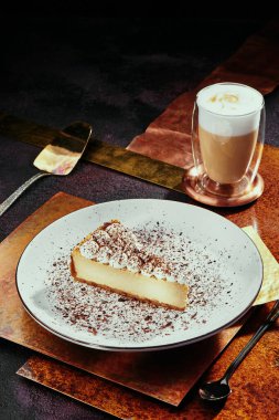 Cheesecake ve kapuçino. Krem kapaklı cheesecake ve bakır kaplamalı beyaz tabakta çikolata parçaları. Kapat.