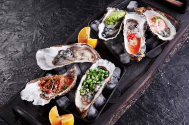 Buzlu ve limonlu tabakta çeşitli taze istiridyeler. Arka planda siyah dokular, üst manzara.