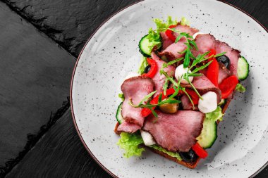 Greek toast with meat slices, tomatoes, mozzarella, olives and lettuce on a white plate isolated on a black stone background. Top view. Space for text