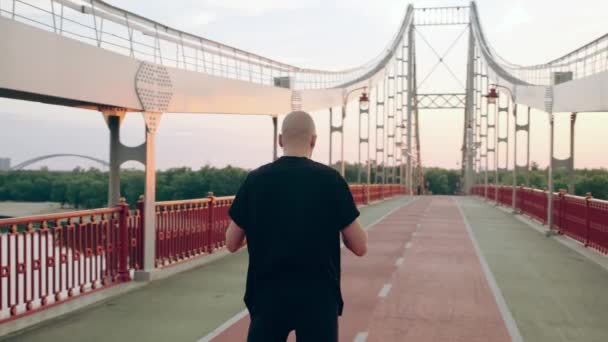 El joven de camiseta negra se pone el auricular y corre por el puente peatonal al amanecer. Movimiento lento — Vídeo de stock