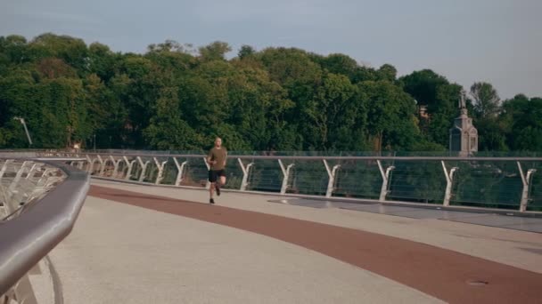 Un joven con uniforme deportivo corre por el puente peatonal al amanecer. Movimiento lento — Vídeos de Stock