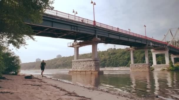 Młody mięśniak biegnie pod mostem w słoneczny dzień. Ćwiczenia serca. Poranne bieganie — Wideo stockowe