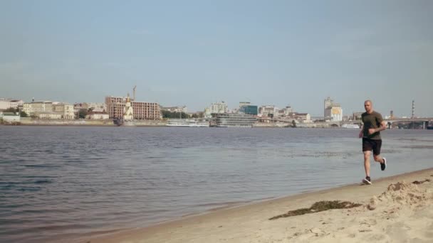 Un joven musculoso corriendo por la playa de la ciudad en un día soleado. Ejercicio cardiovascular. Mañana corriendo — Vídeo de stock