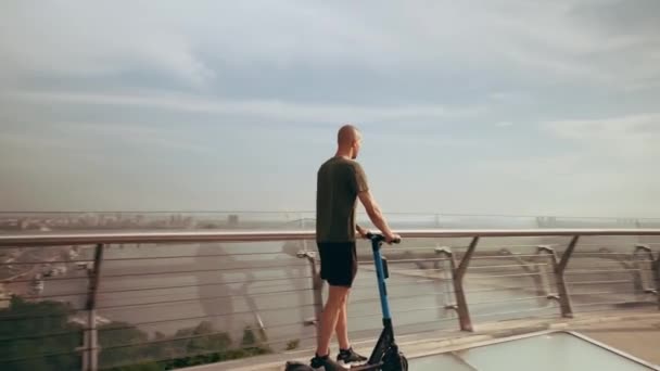 Jovem moderno monta uma scooter elétrica através da ponte pedonal com vista para a cidade — Vídeo de Stock