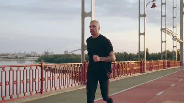 Un joven con uniforme deportivo negro corre por el puente peatonal al amanecer. Movimiento lento — Vídeos de Stock