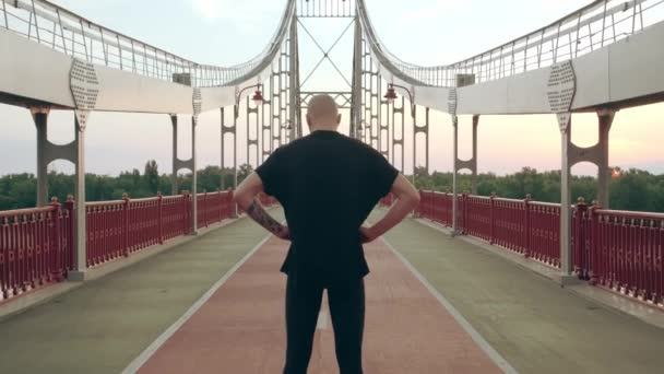 Młody biały człowiek w czarny sport mundur robi rozgrzewkę szyi przed uruchomić most dla pieszych o świcie — Wideo stockowe