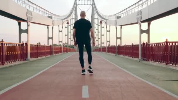 Чоловік бігун в чорній спортивній формі розтягування ніг перед спортом на пішохідному мосту на світанку — стокове відео