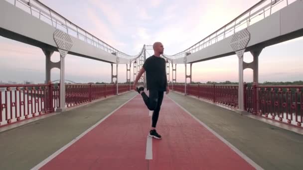 Hombre corredor en negro deporte uniforme estiramiento piernas antes de los deportes en puente peatonal al amanecer — Vídeo de stock