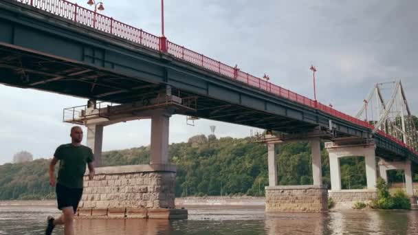 Um jovem musculoso a correr debaixo da ponte num dia ensolarado. Exercício cardiológico. De manhã. — Vídeo de Stock