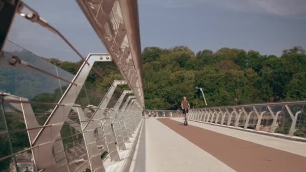 Joven moderno monta un scooter eléctrico a través del puente peatonal con vistas a la ciudad — Vídeos de Stock