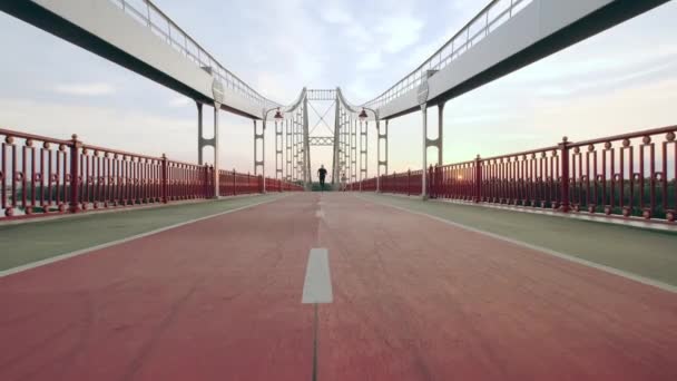 Un joven con uniforme deportivo negro corre por el puente peatonal al amanecer. Movimiento lento — Vídeo de stock