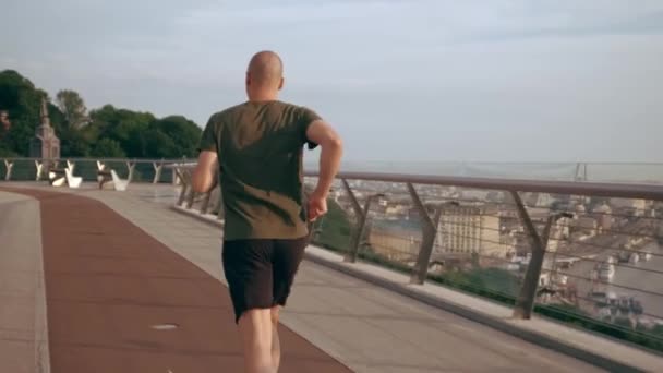 Mladý muž ve sportovní uniformě běhá za úsvitu po mostě pro pěší. Zpomalený pohyb — Stock video