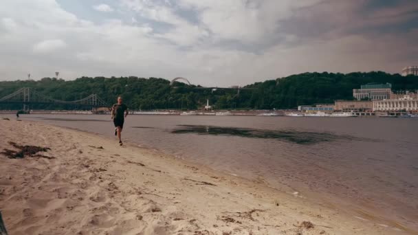 Un joven musculoso corriendo por la playa de la ciudad en un día soleado. Ejercicio cardiovascular. Mañana corriendo — Vídeo de stock