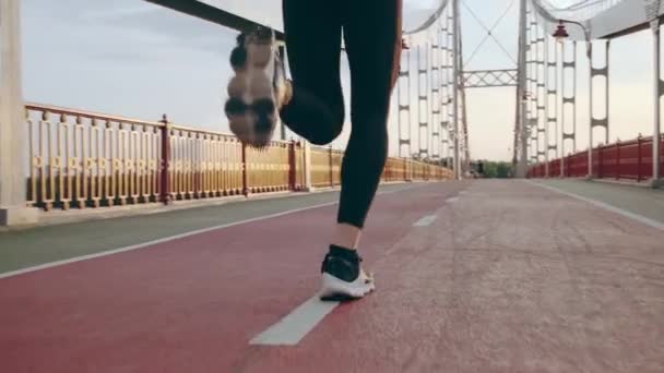 Un joven con uniforme deportivo negro corre por el puente peatonal al amanecer. Movimiento lento — Vídeo de stock