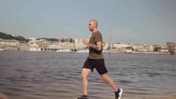 Jovem musculoso correndo na praia da cidade em um dia ensolarado. Exercício cardiológico. De manhã. — Vídeo de Stock