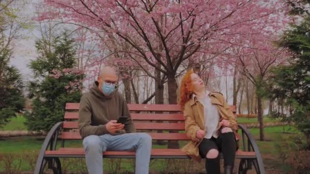Europäischer Mann mit Maske und Frau ohne Maske sitzen auf Bank im Park. Frau hustet ansteckend. Coronavirus-Epidemie — Stockvideo