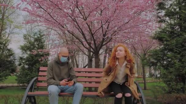 Европейский мужчина в маске и женщина без маски сидят на скамейке в парке. Женщина кашляет заразно. Эпидемия коронавируса — стоковое видео