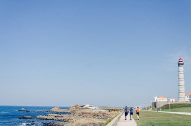 Portekiz 'deki Atlantik Okyanusu kıyısındaki Lea fenerine doğru manzaralı bir kıyı yolunda yürüyen turistler