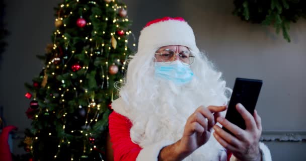 Santa claus com máscara médica está sentado na sala de estar e conversando por smartphone com vídeo chat. — Vídeo de Stock