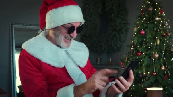 Hombre alegre en gafas negras en traje de Santa Claus está sentado en la sala de estar y charlando por teléfono inteligente con chat de vídeo. — Vídeo de stock