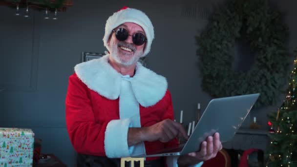 Wesoły mężczyzna w czarnych okularach w stroju Świętego Mikołaja siedzi w salonie i rozmawia przez laptopa z czatem wideo. — Wideo stockowe