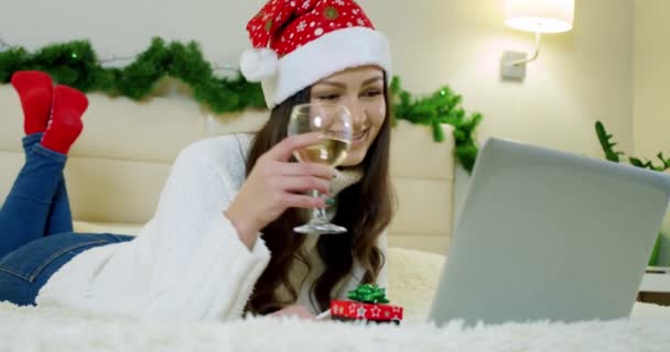 Vacaciones Navidad interior concepto de invierno. mujer joven en un acogedor dormitorio con computadora portátil tumbada en la cama en casa y sosteniendo una copa de champán. Movimiento lento — Vídeo de stock