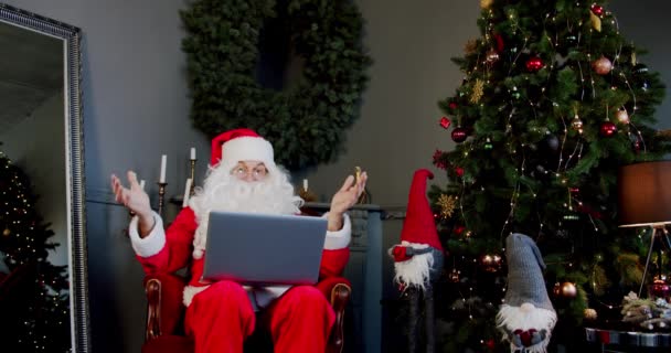 Mężczyzna w stroju Świętego Mikołaja siedzi w salonie i rozmawia przez laptopa z czatem wideo. Szeroki strzał. — Wideo stockowe