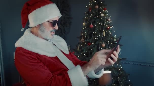 Veselý Santa Claus v černých brýlích sedí v obývacím pokoji a chatuje u smartphonu s video chat. — Stock video