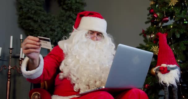 Санта Клаус, сидящий в кресле с ноутбуком на коленях, делает онлайн-покупки с карточкой. Закрыть — стоковое видео