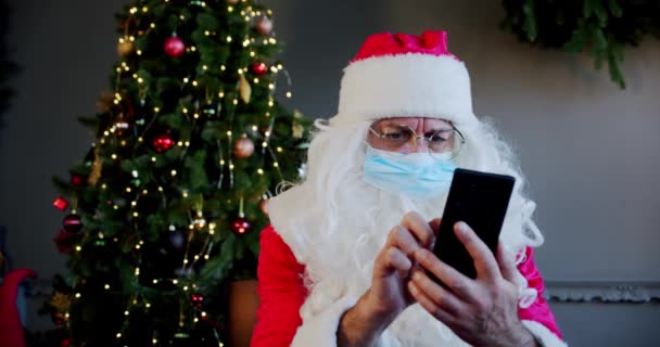 Santa claus with medical mask in living room and chatting by smartphone with video chat. Close up — Stock Video