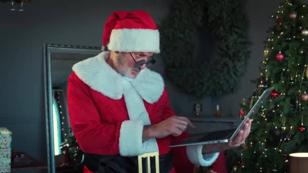 Wesoły człowiek w czarnych okularach w stroju Świętego Mikołaja siedzi w salonie i rozmawia przez laptopa z czatem wideo. Zwolniony ruch — Wideo stockowe