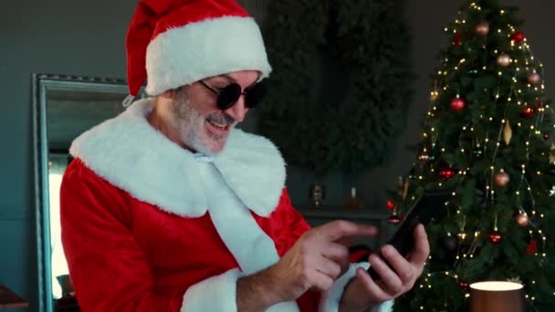 Veselý muž v černých brýlích v obleku Santa Claus sedí v obývacím pokoji a chatuje smartphonem s video chatem. — Stock video