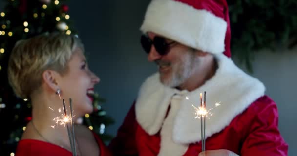 Счастливая пара женщина и мужчина в черных очках и одежда Санта-Клауса держа бенгальские огни на новогодней части. Медленное движение — стоковое видео
