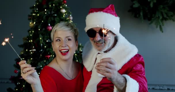 Mujer feliz pareja y el hombre en gafas negras y ropa de Santa Claus celebración de bengalas en la parte de año nuevo. Cámara lenta Primer plano — Vídeo de stock