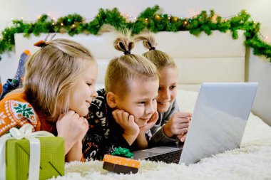 Yılbaşı konsepti, çocuklar Noel kıyafetleriyle kanepede yatar ve bir dizüstü bilgisayarla internetten arama yaparlar..