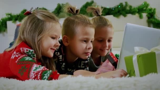 Concept Nouvel An, les enfants s'allongent sur le canapé en vêtements de Noël, et faire un appel en ligne sur un ordinateur portable. Fermer les enfants — Video