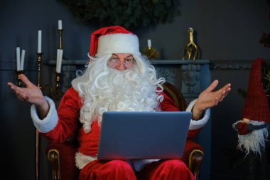 Noel Baba kostümlü adam oturma odasında oturmuş bilgisayarın başında görüntülü sohbet ediyor..