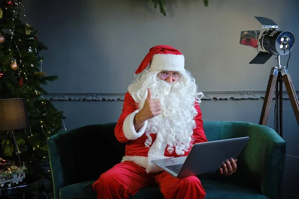 Noel Baba kostümlü adam oturma odasında oturmuş bilgisayarın başında görüntülü sohbet ediyor ve parmağını kaldırıyor.