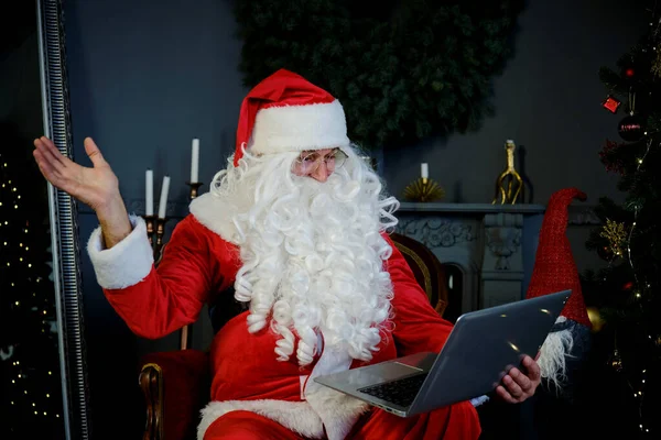 Mężczyzna w stroju Świętego Mikołaja siedzi w salonie i rozmawia przez laptopa z czatem wideo i macha ręką. Zamknij się. — Zdjęcie stockowe