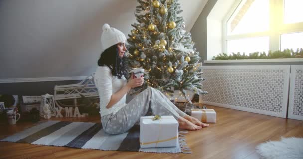 Girl in blue pajamas sits on the floor and holds a cup near the Christmas tree with gifts. New year concept — Stock Video