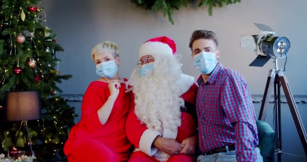 Jovem casal usando máscaras faciais e chapéu de Papai Noel sentado em casa. Tiro largo — Vídeo de Stock